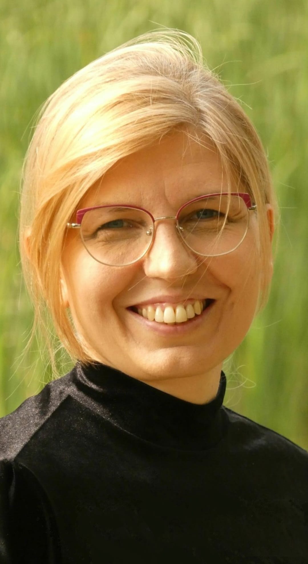 Uśmiechnięta profesor Agnieszka Korgul w dużych okularach i czarnym golfie, na tle zieleni.